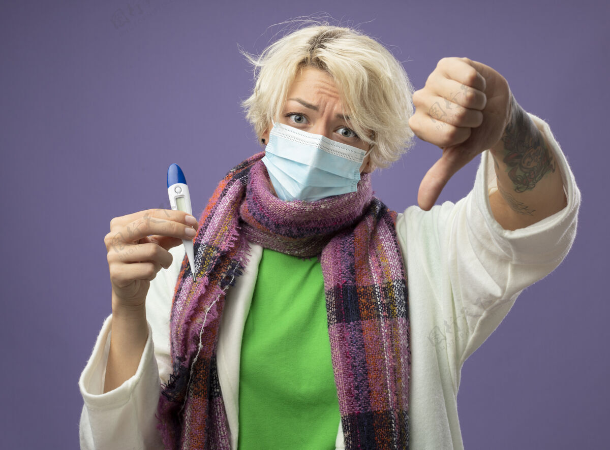 生病不快乐的生病的不健康的女人 短头发 戴着暖和的围巾 戴着面罩 拿着温度计 拇指朝下竖立在紫色的背景上短裤抱面具