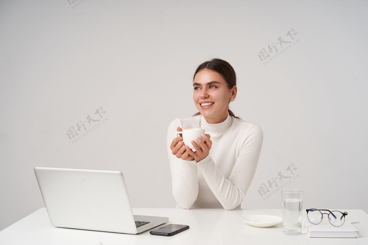 手机室内拍摄的是一位穿着正装 有魅力的黑发年轻女子 在办公室里拿着笔记本电脑喝着咖啡 隔着白墙Mua头发女士