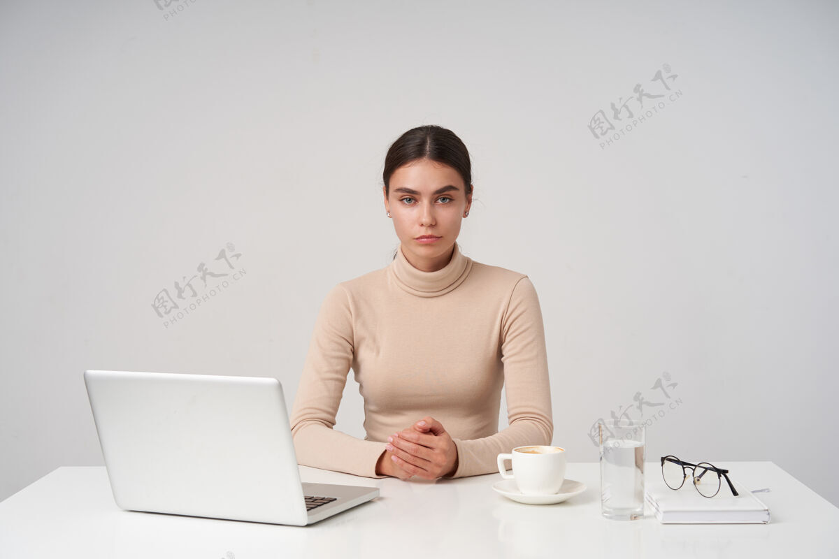 优雅严肃的年轻蓝眼睛黑发女性在现代办公室工作 带着笔记本电脑 看的时候嘴唇合拢 穿着正式的衣服 在白色的墙上摆姿势年轻爱情眼镜