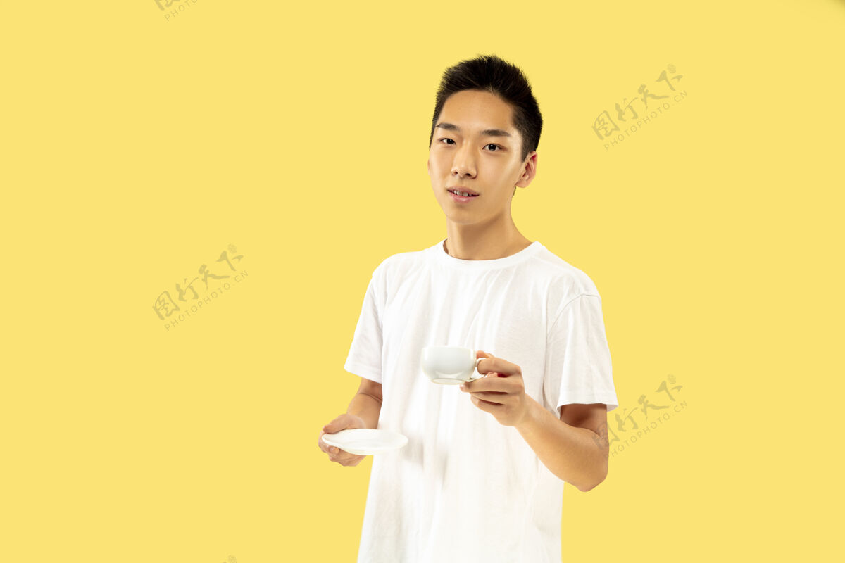 成功黄色摄影棚背景上的韩国年轻人半身像穿白衬衫的男模喝咖啡 感觉快乐人类情感的概念 面部表情正面视图流行色彩肖像亚洲人衬衫