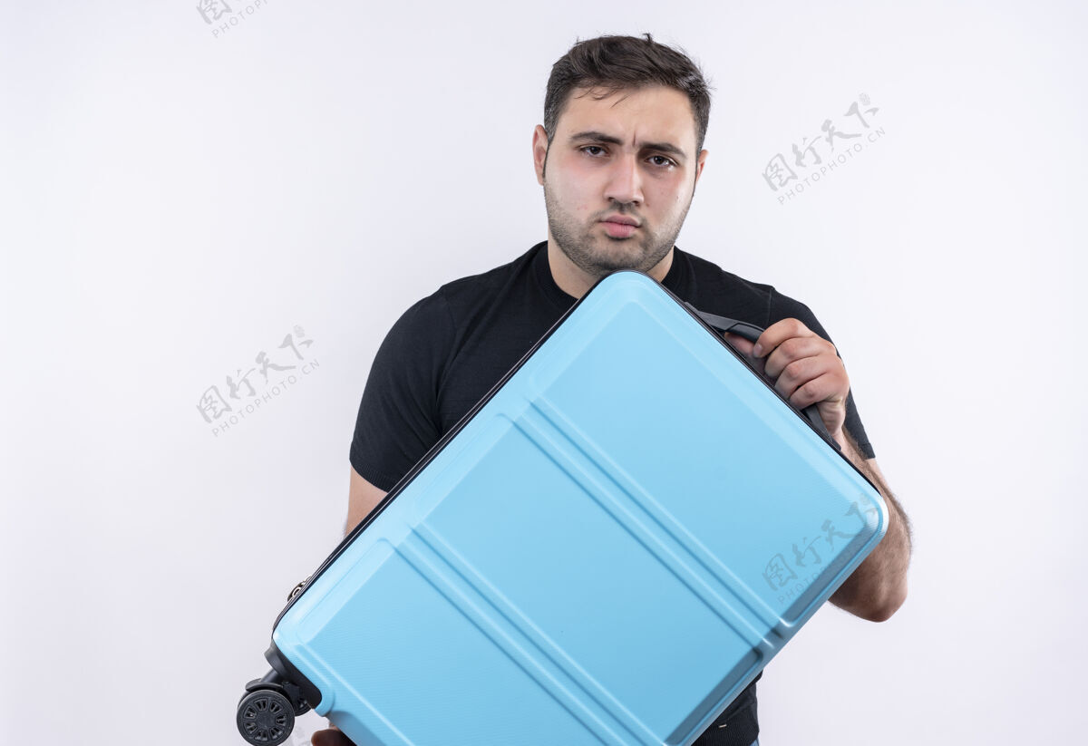 抱着一个穿着黑色t恤的年轻旅行者站在白色的墙上 手里拿着手提箱 严肃地皱着眉头旅行摆姿势假日