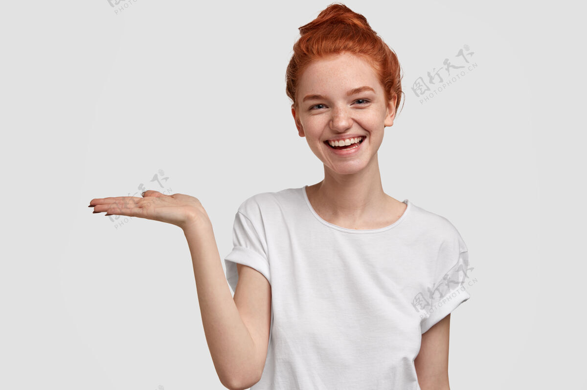 内容满意可爱的欧洲女孩用姜红色的发髻撑起临摹空间 掌心抬高 靠着白色的墙壁做广告 有俏皮的表情显示高兴成人