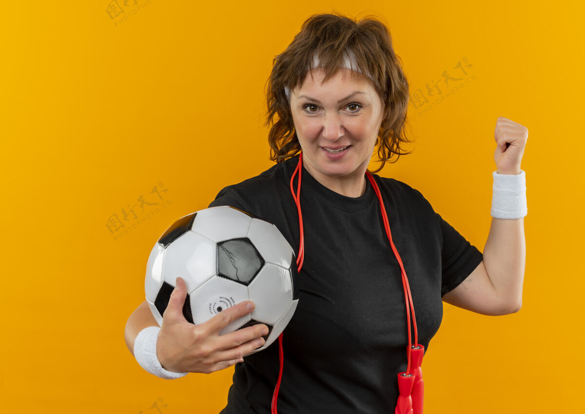 女身穿黑色t恤的中年运动女性 头箍举着足球 举起拳头 快乐而积极 冠军概念站在橙色的墙上赢家人中间