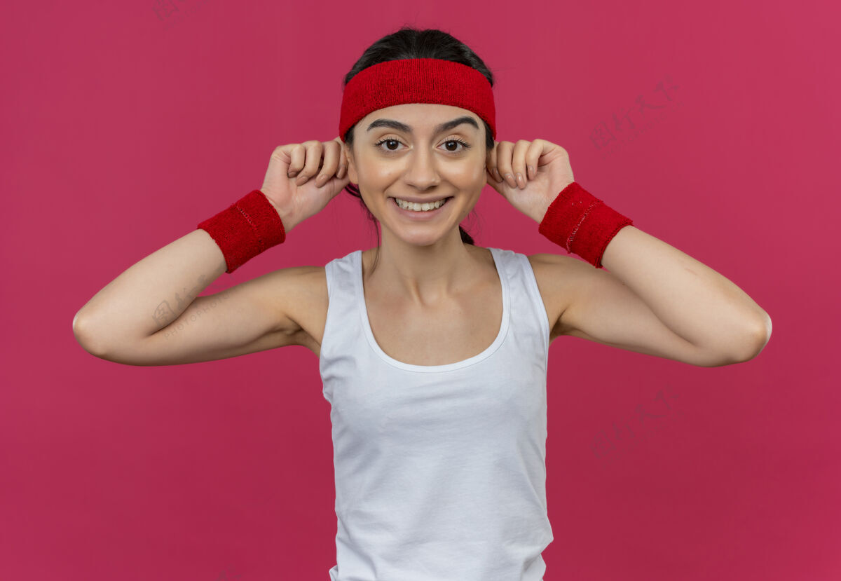 女人身穿运动服 头箍贴着耳朵 脸上挂着笑容的年轻健身女士站在粉色的墙上运动年轻健身