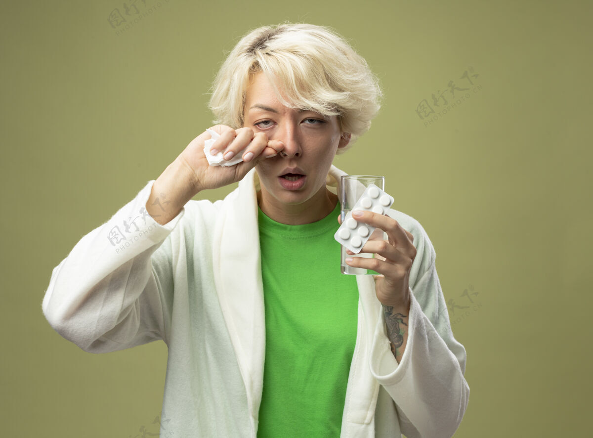 湿巾一位短发妇女生病了 她拿着一杯水 拿着药丸 站在灯光下用餐巾擦鼻子 感觉不舒服疾病鼻子药片