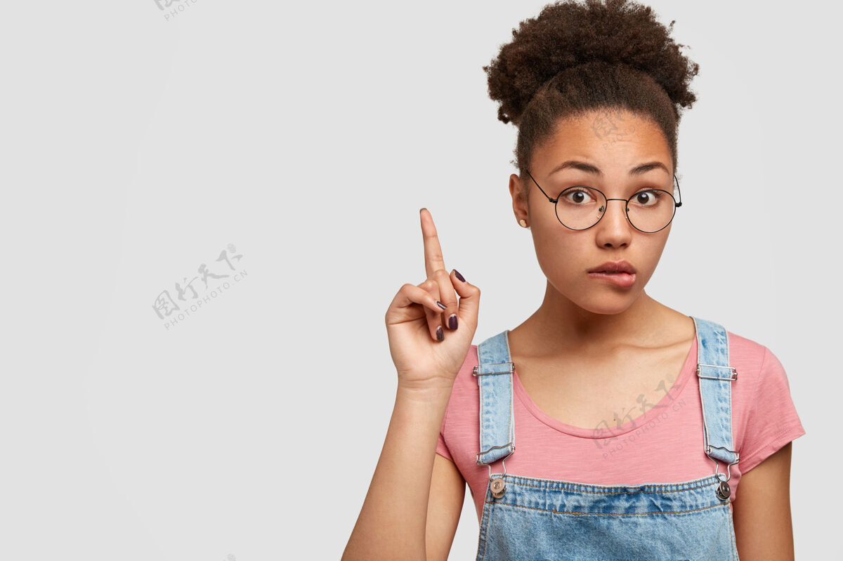 女人惊讶不解的非裔美国女士咬下唇 食指向上指 建议抬头 神色不定 戴眼镜 靠着白墙 抄空格指向时尚时尚