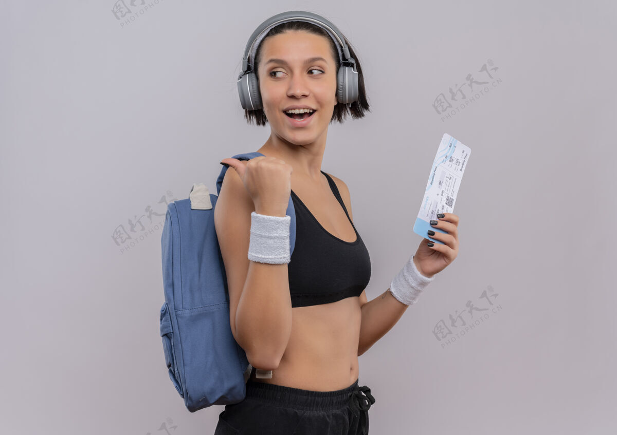 拿着穿着运动服的年轻健身女士头戴耳机背着背包拿着机票看着一边用拇指指着后面站在白色的墙上票女运动装