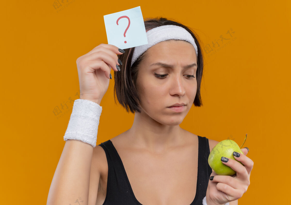 抱着身穿运动服的年轻健身女士 头上戴着带问号的提醒纸 站在橙色的墙上 绿苹果带着沉思的表情看着苹果女性纸市民