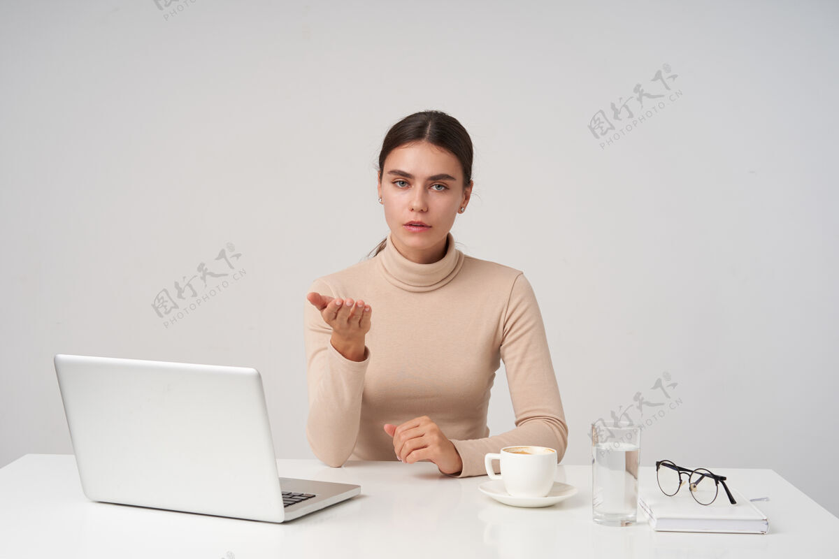 姿势室内照片 年轻漂亮的黑发女士穿着米色马球衫 拿着笔记本电脑和咖啡坐在桌边 困惑地举起手掌 困惑地看着相机笔记本电脑心情肖像