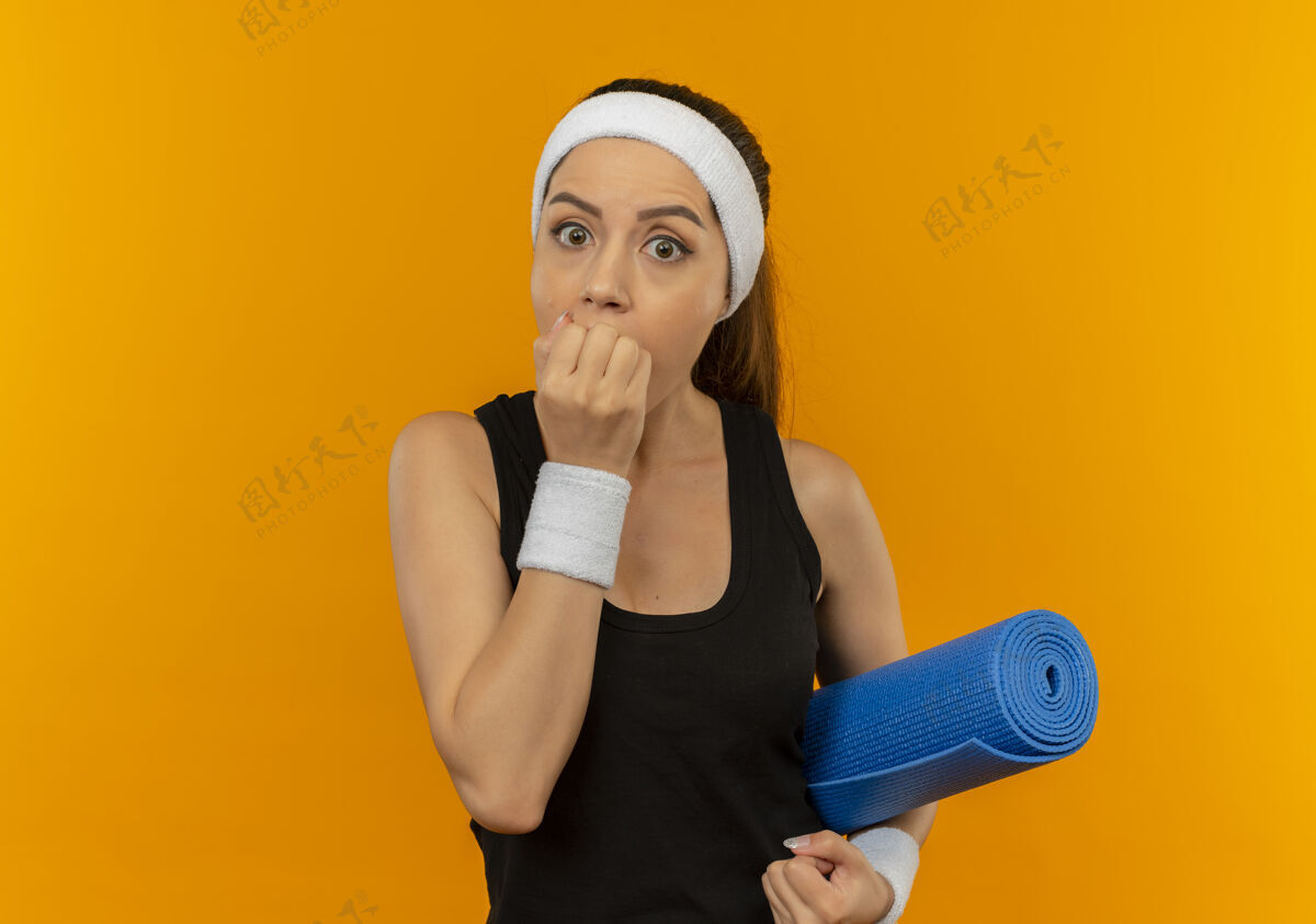 头带身穿运动服的年轻健身女士 头戴瑜伽垫 手捂着嘴站在橙色的墙上站着瑜伽运动