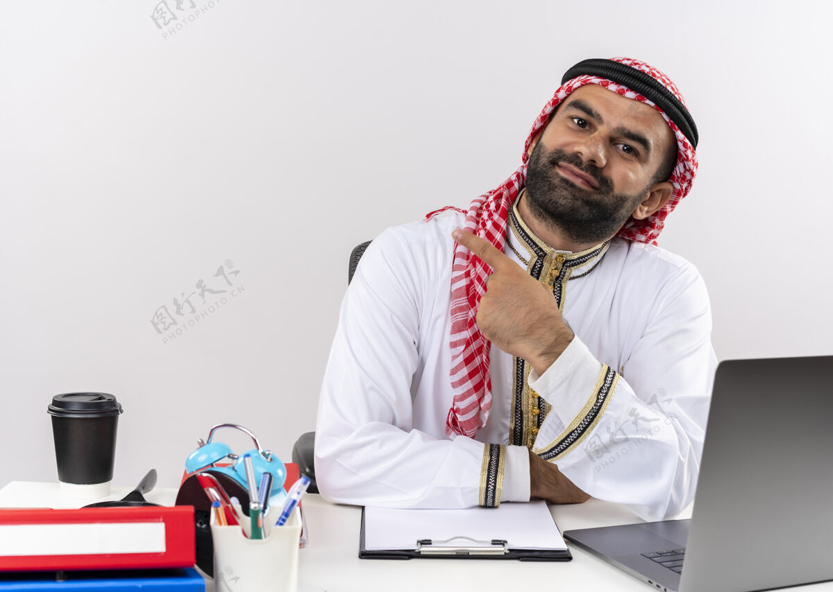 手指身着传统服装的阿拉伯商人坐在桌旁 用食指指着左边的笔记本电脑 看上去自信地在办公室工作电脑左笔记本电脑