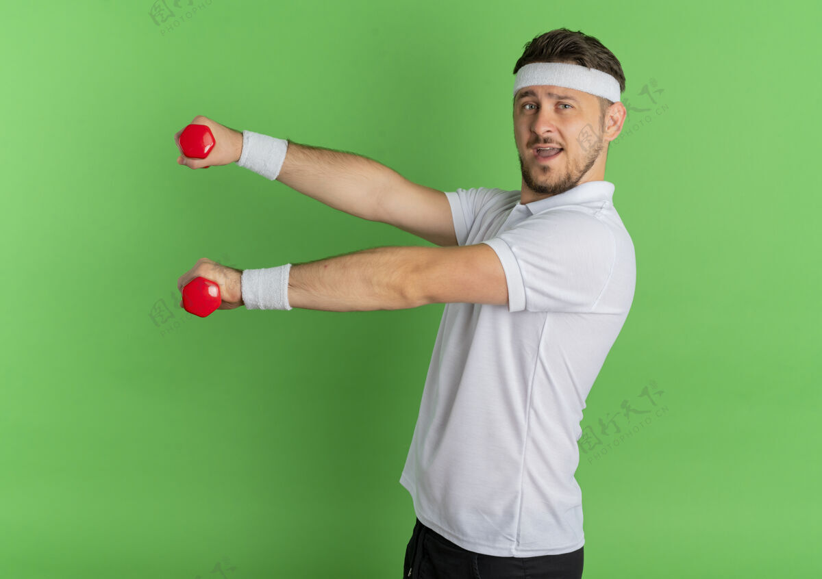 年轻身穿白衬衫 头箍 手持哑铃的年轻健身男子站在绿色的墙壁上快乐而积极地锻炼身体人衬衫姿势