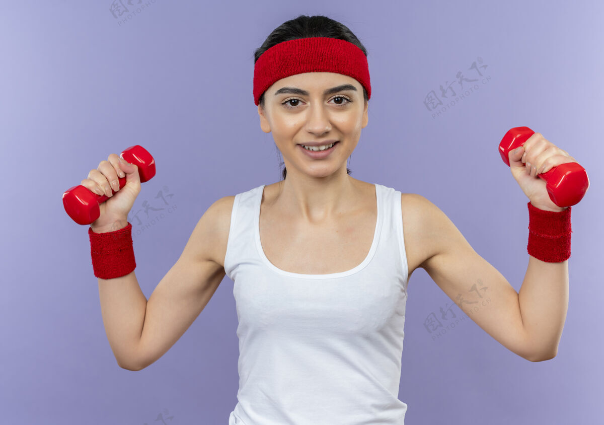 哑铃身穿运动服 头箍 双手举着两个哑铃 面带微笑的年轻健身女孩站在紫色的墙上做运动运动装欢呼举起