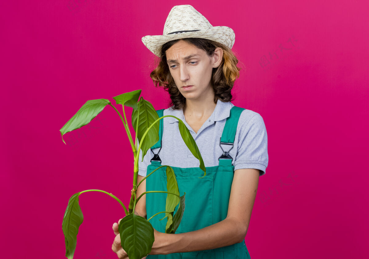 男人年轻的园丁穿着连体衣戴着帽子拿着植物严肃地看着它穿严肃脸