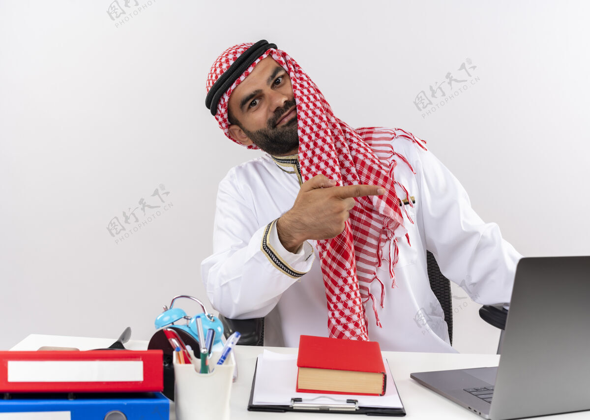 笔记本电脑身着传统服装的阿拉伯商人坐在桌旁 手里拿着笔记本电脑 微笑着 自信地用手指着办公室工作的一侧手指工作商人