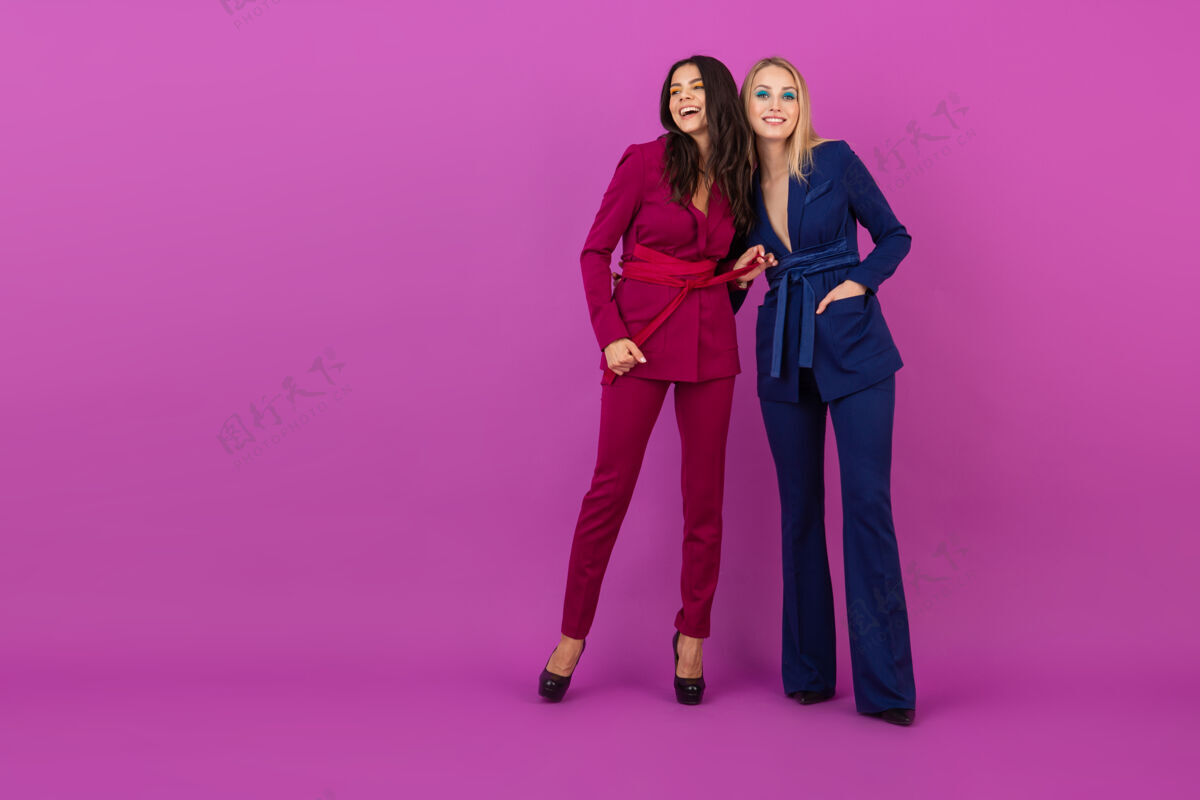 华丽高级时尚风格两位微笑迷人的女士在紫罗兰色的墙壁上穿着时尚多彩的紫色和蓝色晚礼服 朋友们一起玩 时尚潮流时尚年轻优雅