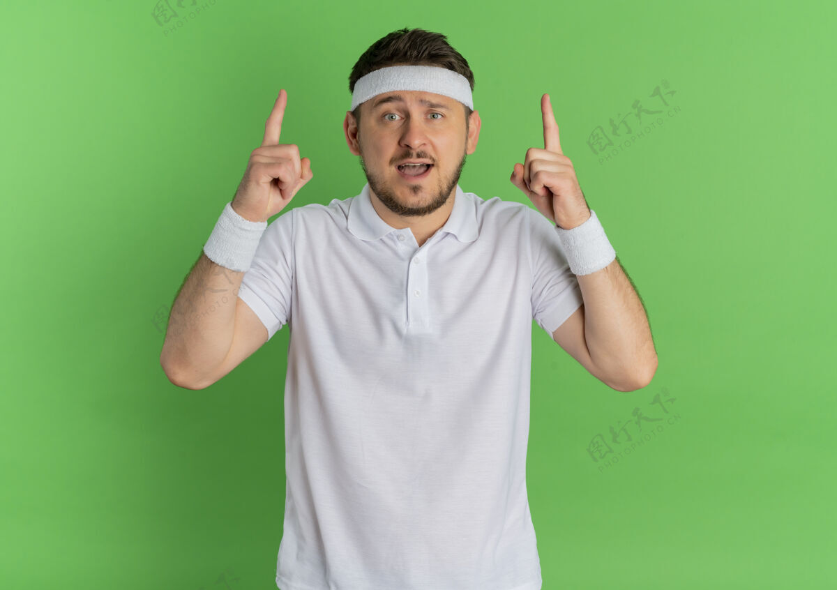 年轻身穿白衬衫 头箍朝上 食指朝前 惊讶地站在绿色墙壁上的年轻健身男子健康运动头带