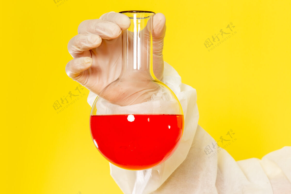 桌子正面近距离观察穿着特殊防护服的男性科学工作者在黄色的桌子上拿着装有红色溶液的烧瓶服装特殊男性