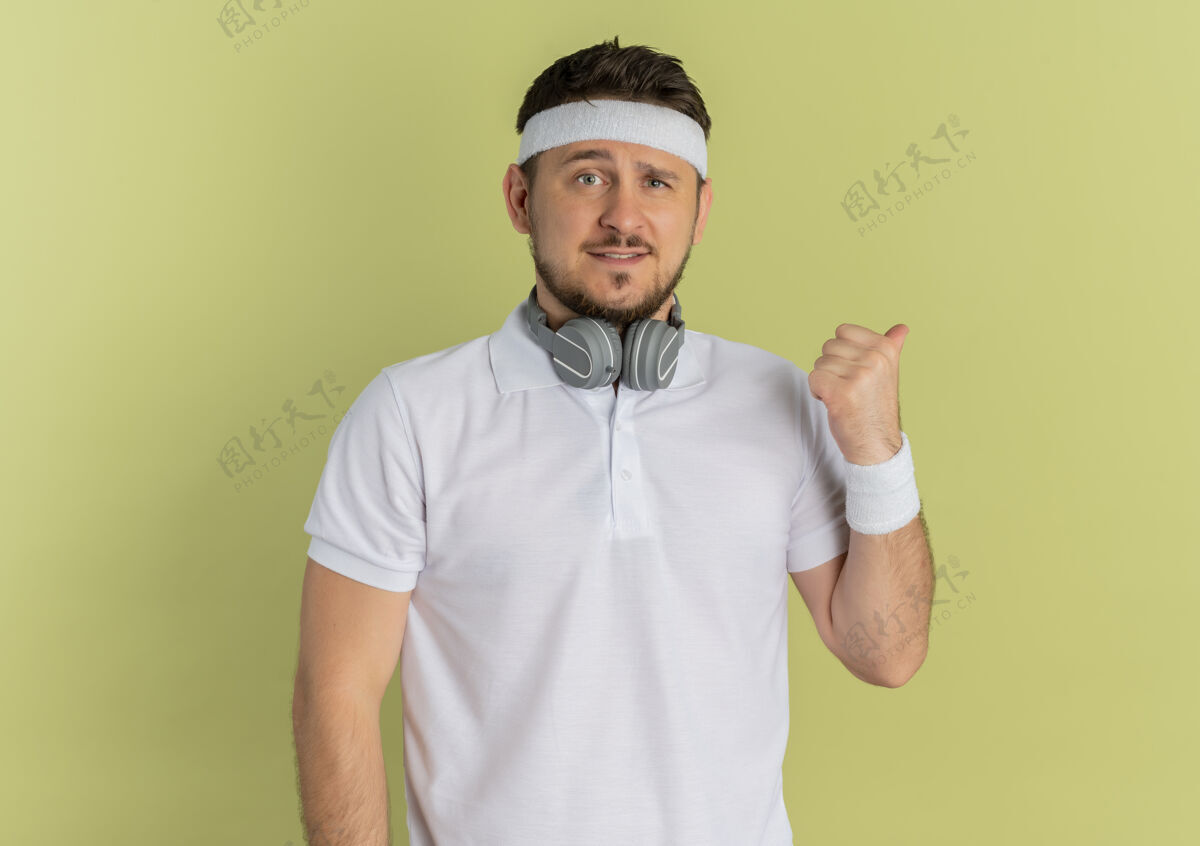 人身穿白衬衫 头箍的年轻健身男子面带微笑 用拇指指着站在橄榄墙上的一侧运动橄榄壁板