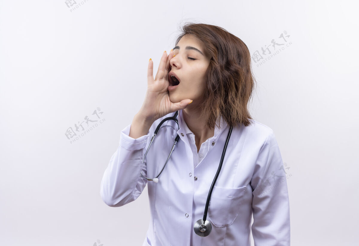 喊叫身穿白大褂 手持听诊器的年轻女医生站在白墙上 用手靠近嘴巴大声喊叫外套站着手