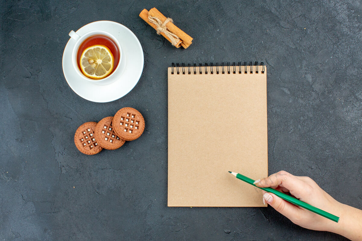 柠檬俯视图一杯茶加柠檬肉桂棒曲奇饼记事本绿色铅笔在女性手上黑色的表面笔记本钢笔书写