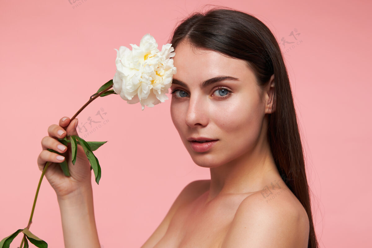 新鲜迷人的 漂亮的女孩的肖像 深色长发和健康的皮肤 用一朵花抚摸着头观看 特写 在粉彩粉色的墙上孤立头发长健康
