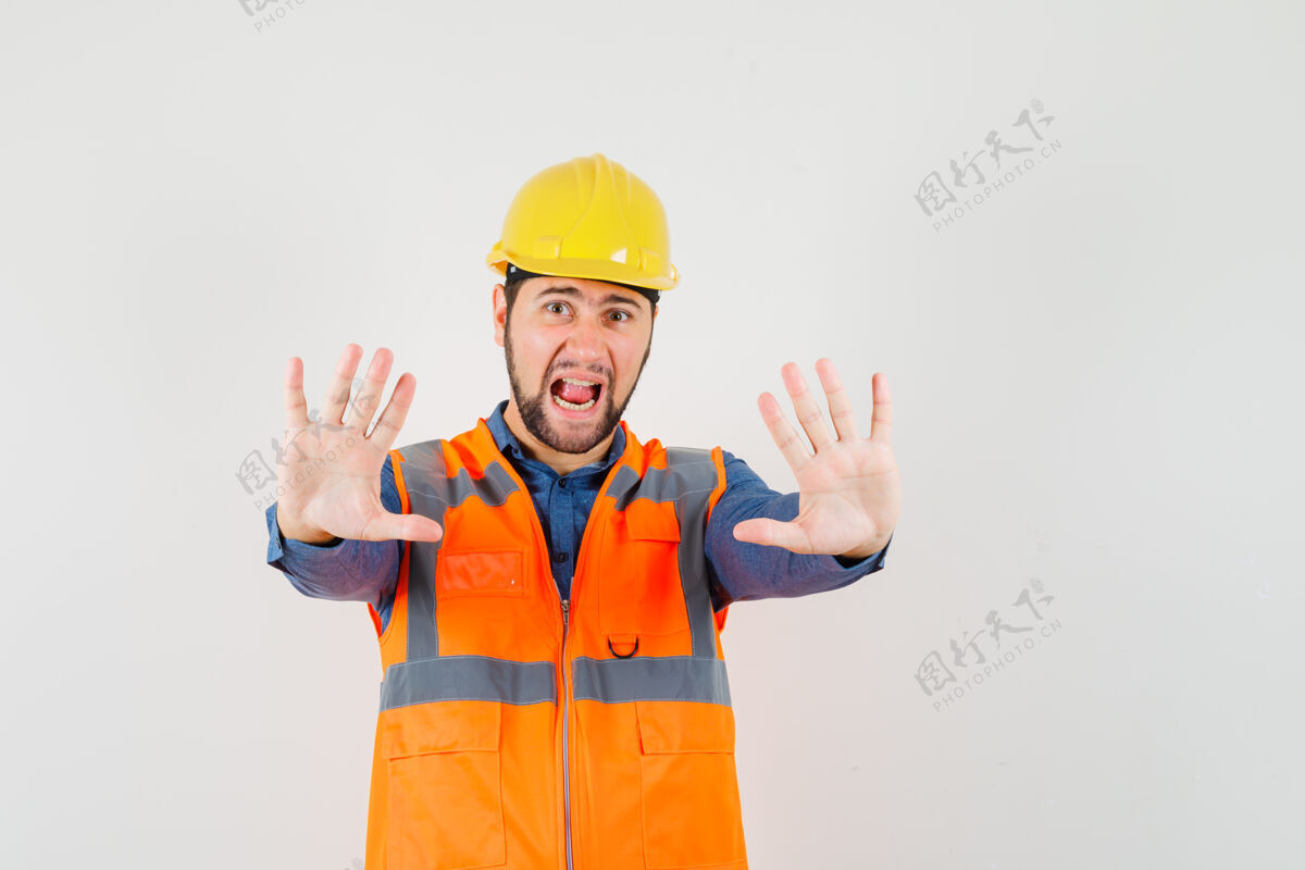 手势年轻的建筑工人穿着衬衫 背心 头盔显示停止手势 同时尖叫 看起来很害怕 前视图制服头盔衬衫