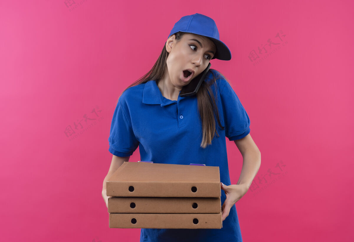 女孩穿着蓝色制服 戴着帽子 拿着一叠比萨饼盒的年轻送货女孩在讲手机时显得很惊讶叠着说话电话