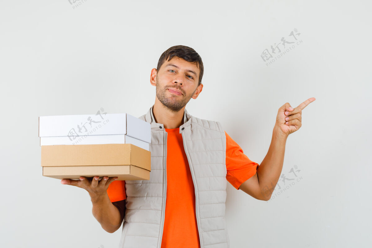 姿势年轻的男性拿着纸板箱 指着远处的t恤衫 夹克衫 看上去很高兴 正面视图男人纸板盒子