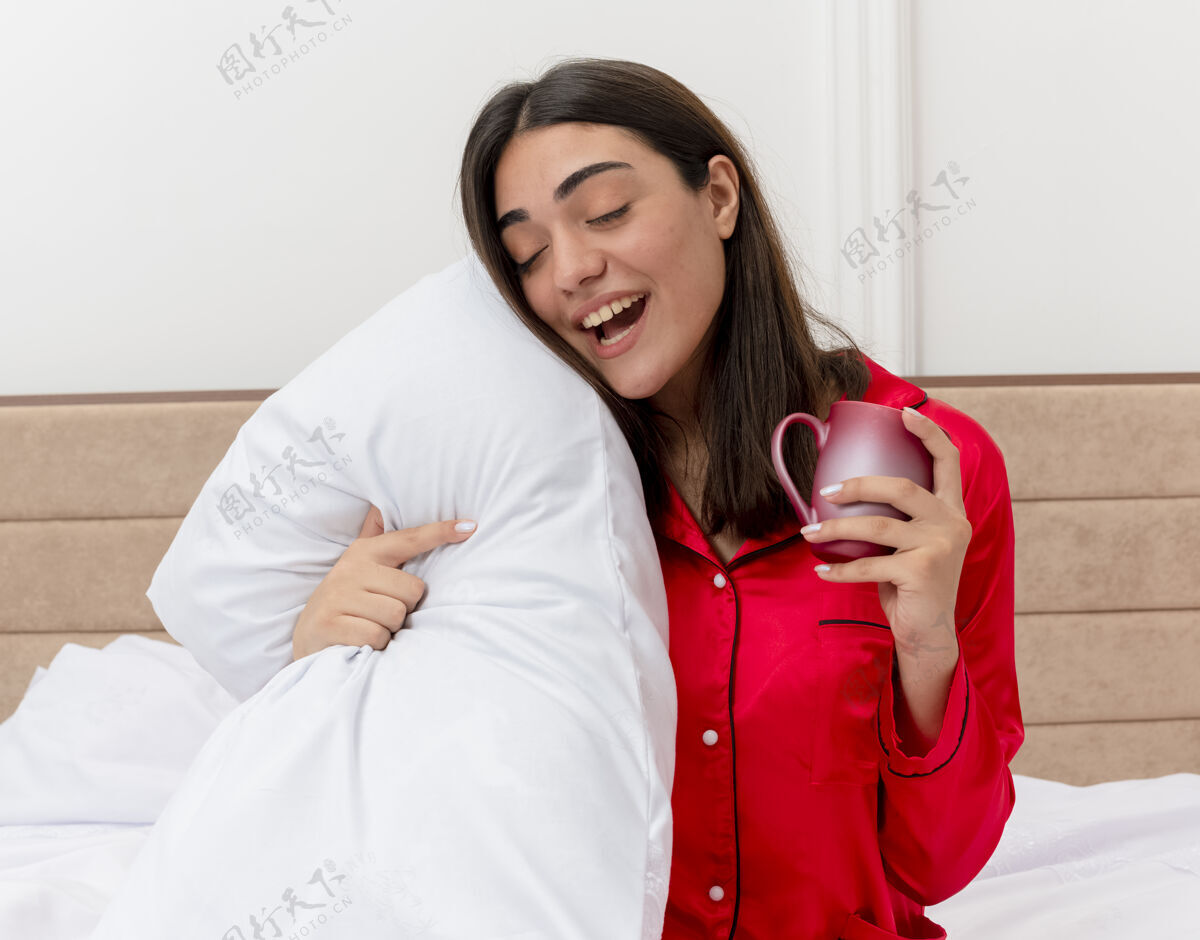 睡衣穿着红色睡衣的年轻美女坐在床上 枕着一个杯子 快乐而积极地放松着 在卧室的室内灯光背景下享受早晨的时光床女人快乐