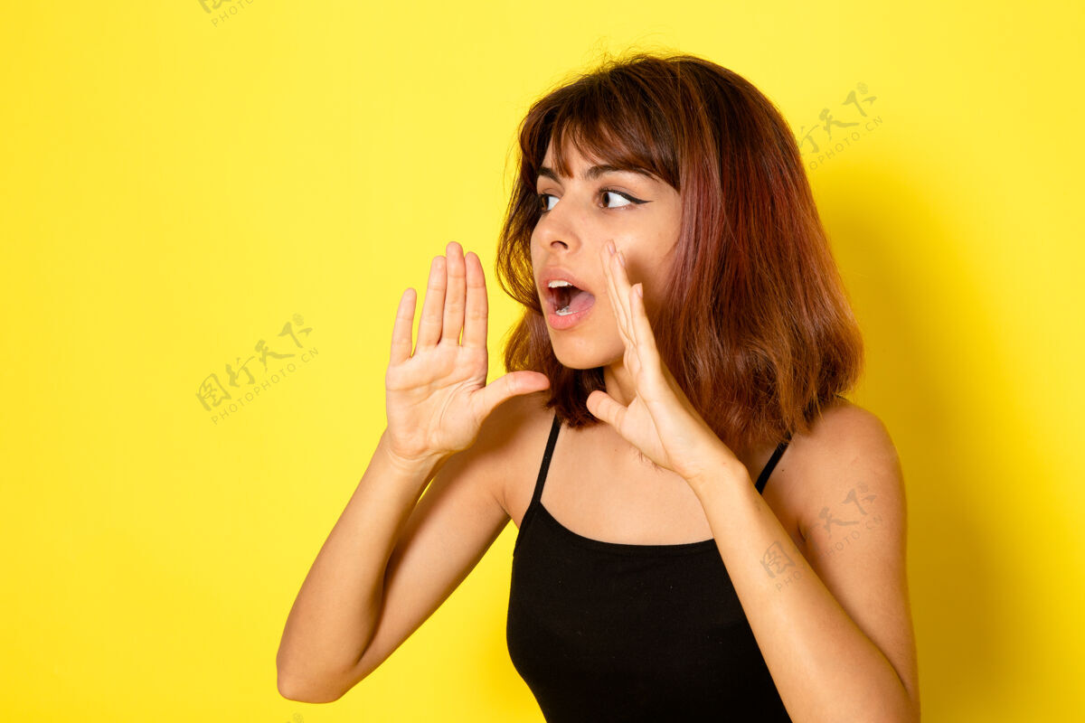 情感身穿黑色衬衫的年轻女性在浅黄色的墙上低声呼唤的正面视图女孩工作耳语