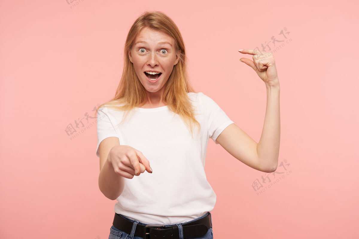 表情年轻兴奋的红头发女性的摄影棚照片 自然妆容 小个子 举手 情绪化地指着镜头 在粉色背景上摆姿势头发女士手指