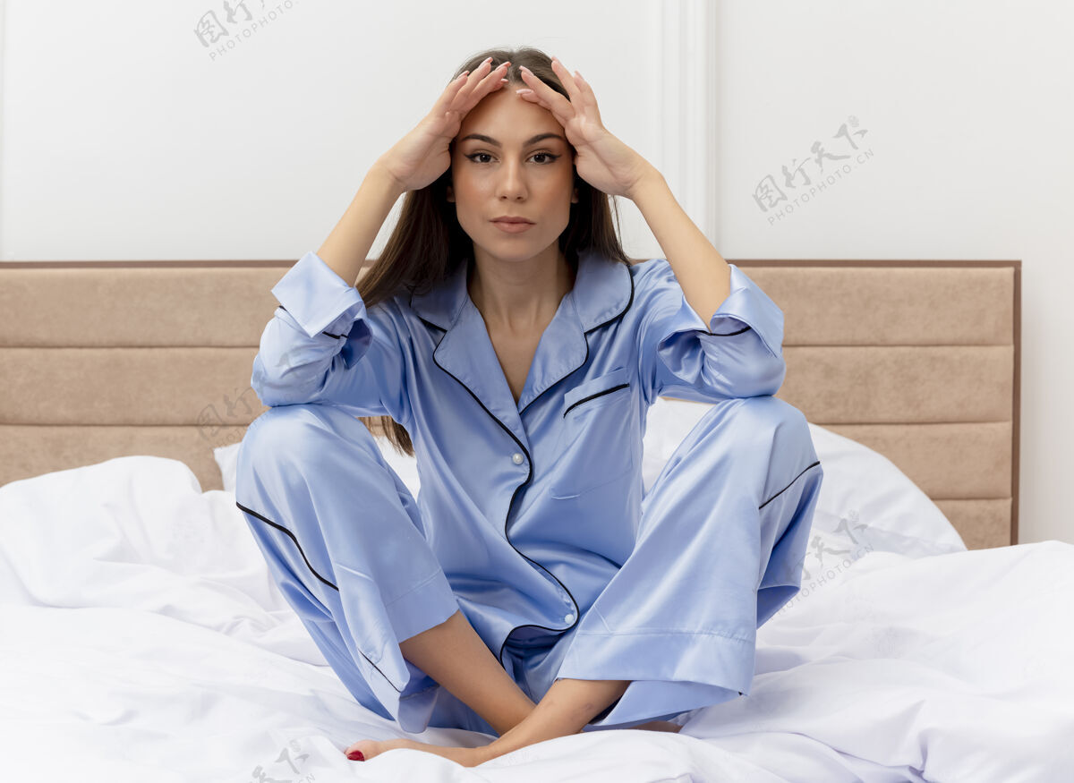 卧室穿着蓝色睡衣的年轻漂亮女人坐在床上看着相机疲惫而无聊地躺在卧室的室内灯光背景下相机室内坐着