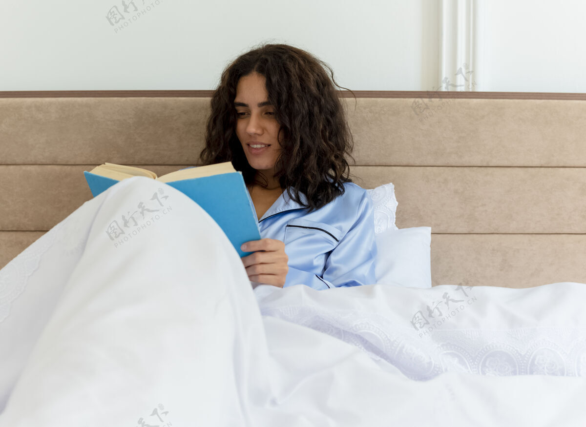 蓝色穿着蓝色睡衣的有趣的年轻漂亮女人坐在床上看书 在卧室的室内灯光背景下享受周末漂亮周末搞笑
