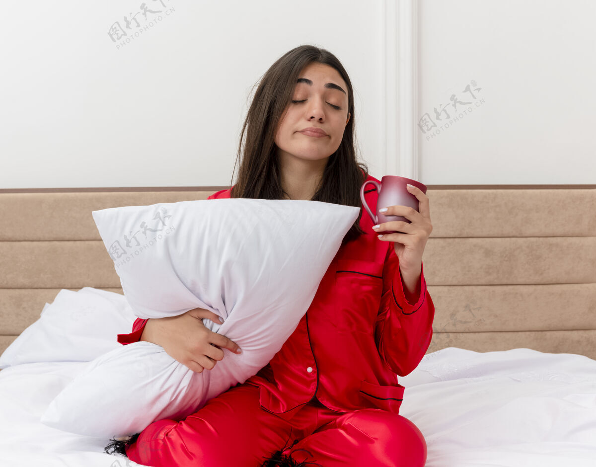 漂亮穿着红色睡衣的年轻美女坐在床上用枕头托着杯子看起来很累感觉早上很累在卧室的室内灯光背景下睡衣卧室枕头