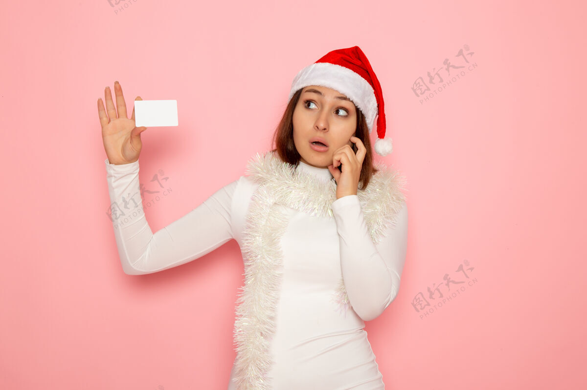 圣诞正面图年轻女子手持白色银行卡 想着粉色墙上圣诞假期新年时尚彩钱肖像美丽风景