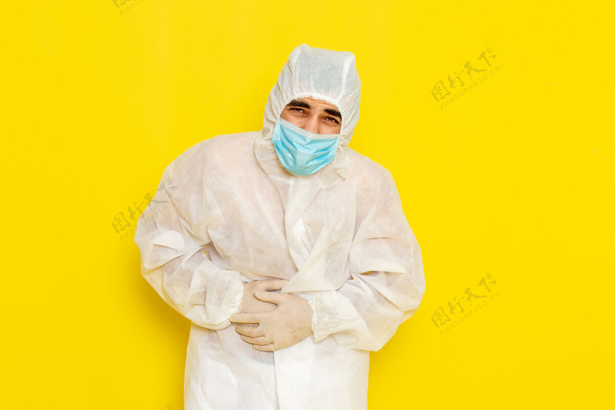 危险身穿白色特殊防护服 浅黄色墙壁上有胃痛口罩的男科学工作者正面图化学胃痛医生