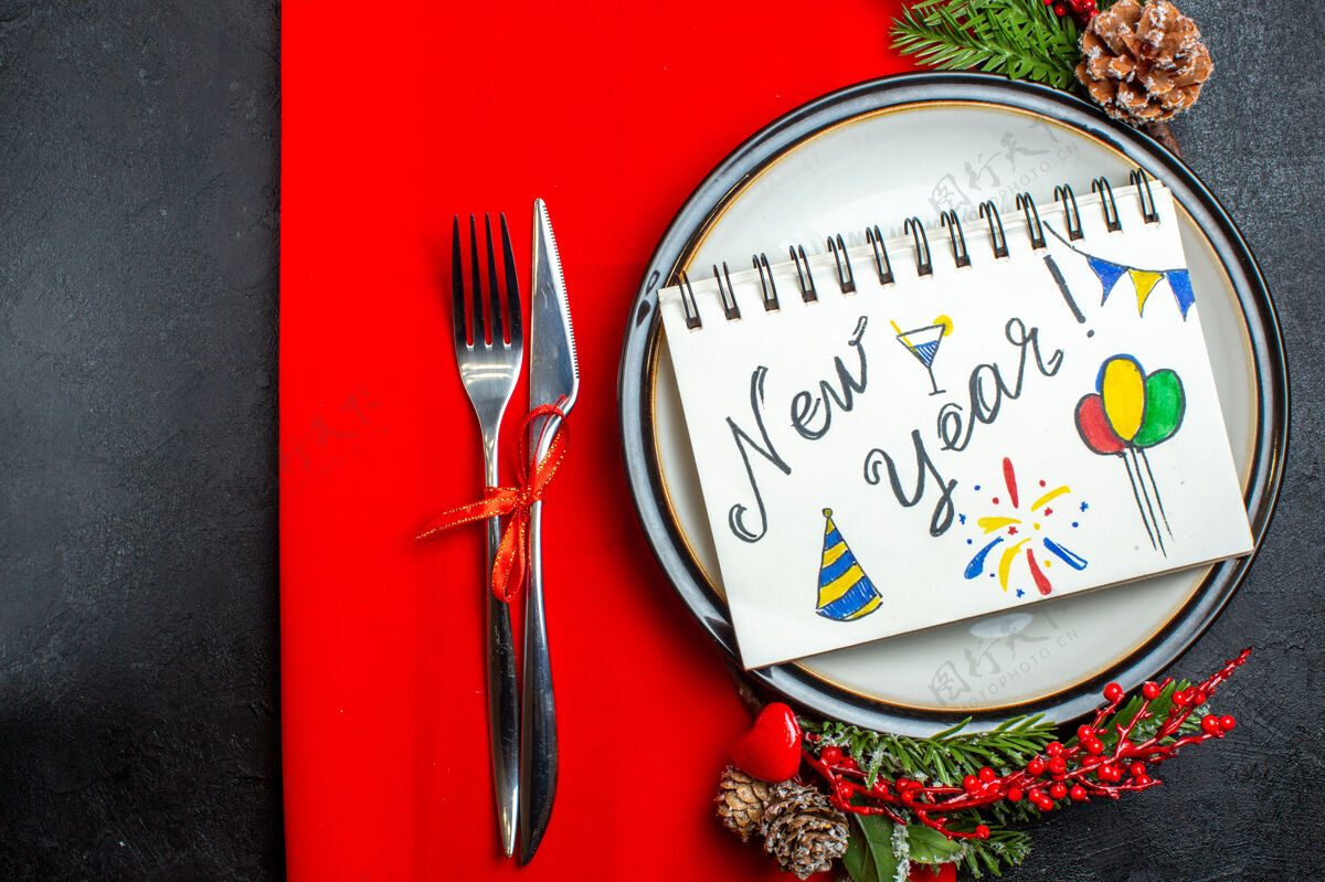 树枝笔记本俯视图 餐盘上有新年文字和图画 装饰配件杉木树枝和餐具放在红色餐巾上叉子画武器