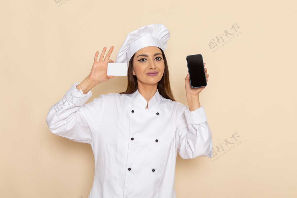 正面身穿白色厨师服的年轻女厨师的正面图 白色墙上拿着白色卡片和电话女性专业电话