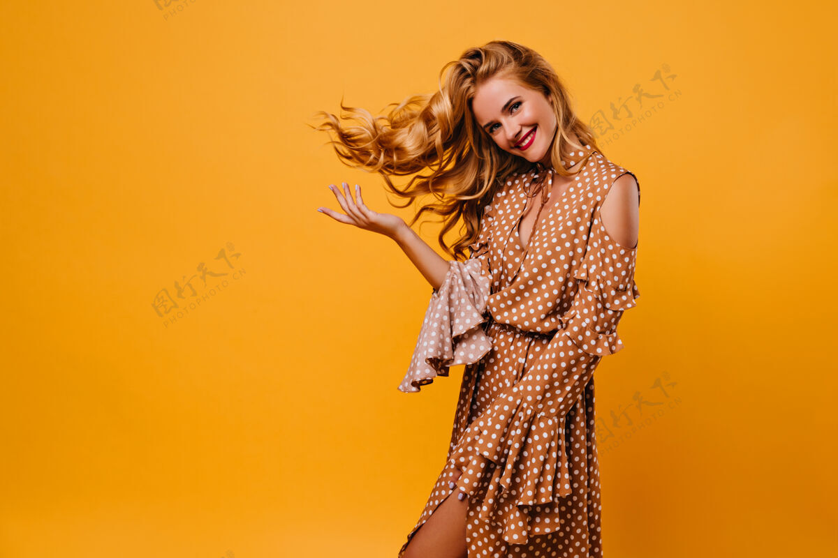 金发快乐的年轻女士 带着迷人的微笑 身着棕色连衣裙室内照片 迷人的金发女郎站在黄色的墙上积极室内年轻