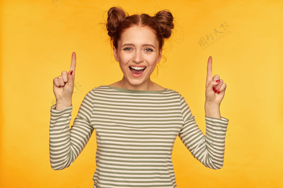 女人十几岁的女孩 快乐的红发女人 带着两个发髻穿着条纹毛衣 面带微笑地看着镜头 快乐地指着复制空间 隔着黄色的墙20多岁面部休闲