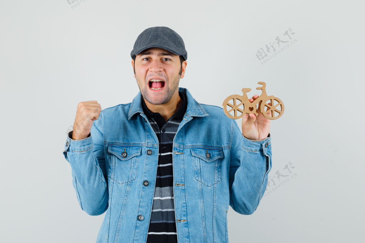 T恤一个穿着t恤 夹克 帽子的年轻人展示着赢家的姿态 手里拿着木制玩具自行车 看上去很幸运手势自行车木头