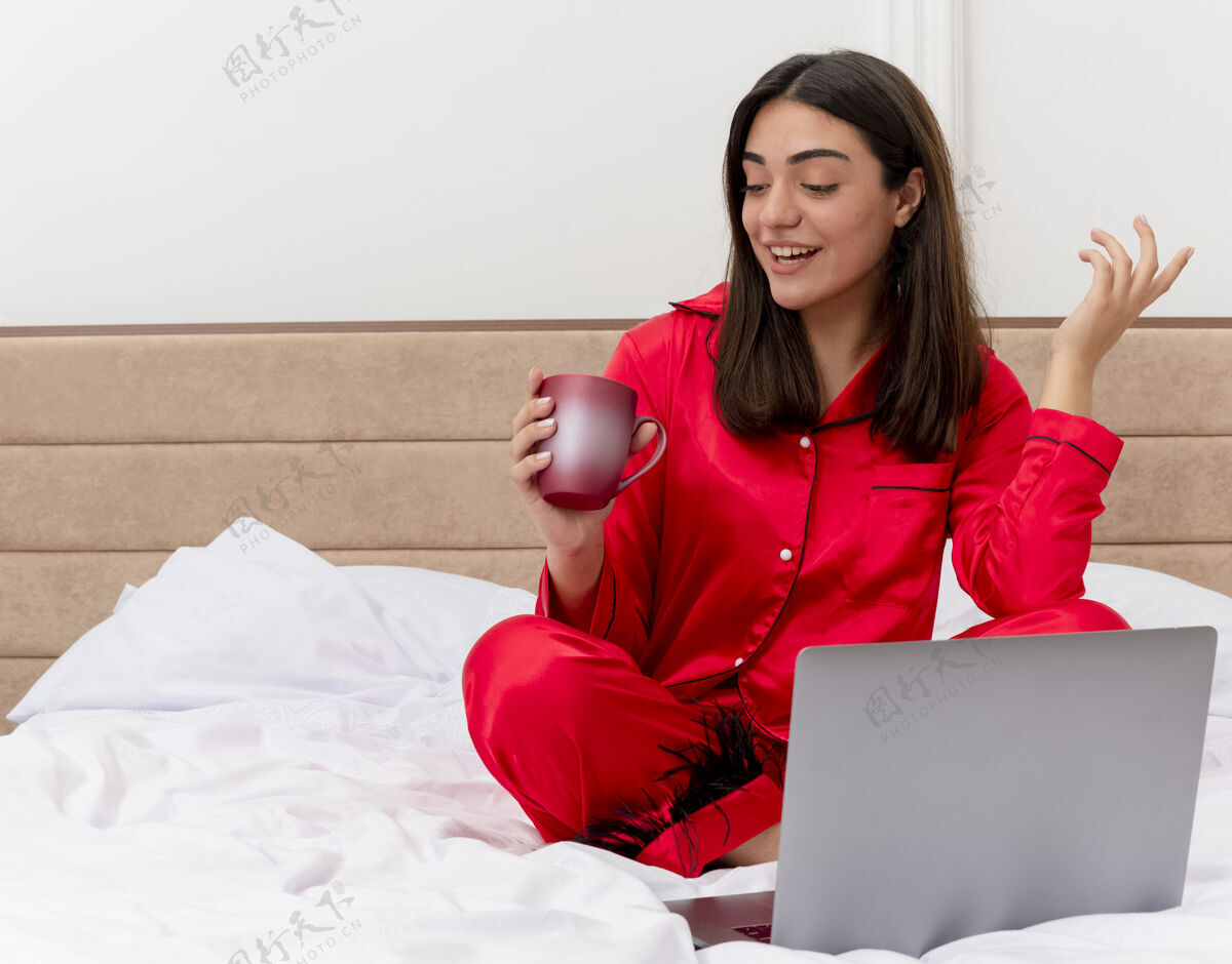笔记本电脑穿着红色睡衣的年轻漂亮女人坐在床上 手里拿着笔记本电脑和一杯咖啡 看着杯子 在卧室的室内灯光背景下开心而积极地微笑着快乐卧室杯子