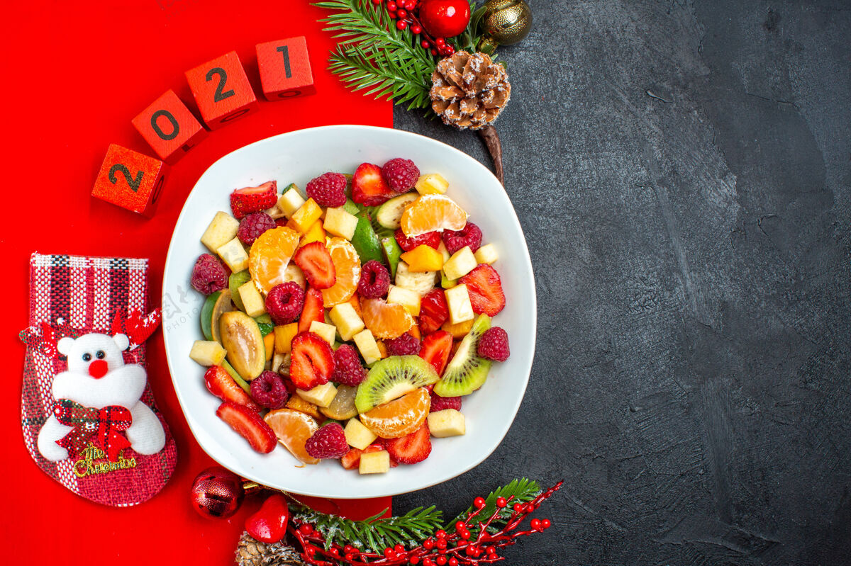 晚餐收集新鲜水果的半张照片放在餐盘装饰配件杉木树枝和数字上 红色餐巾放在深色背景的右侧冷杉圣诞节胡椒