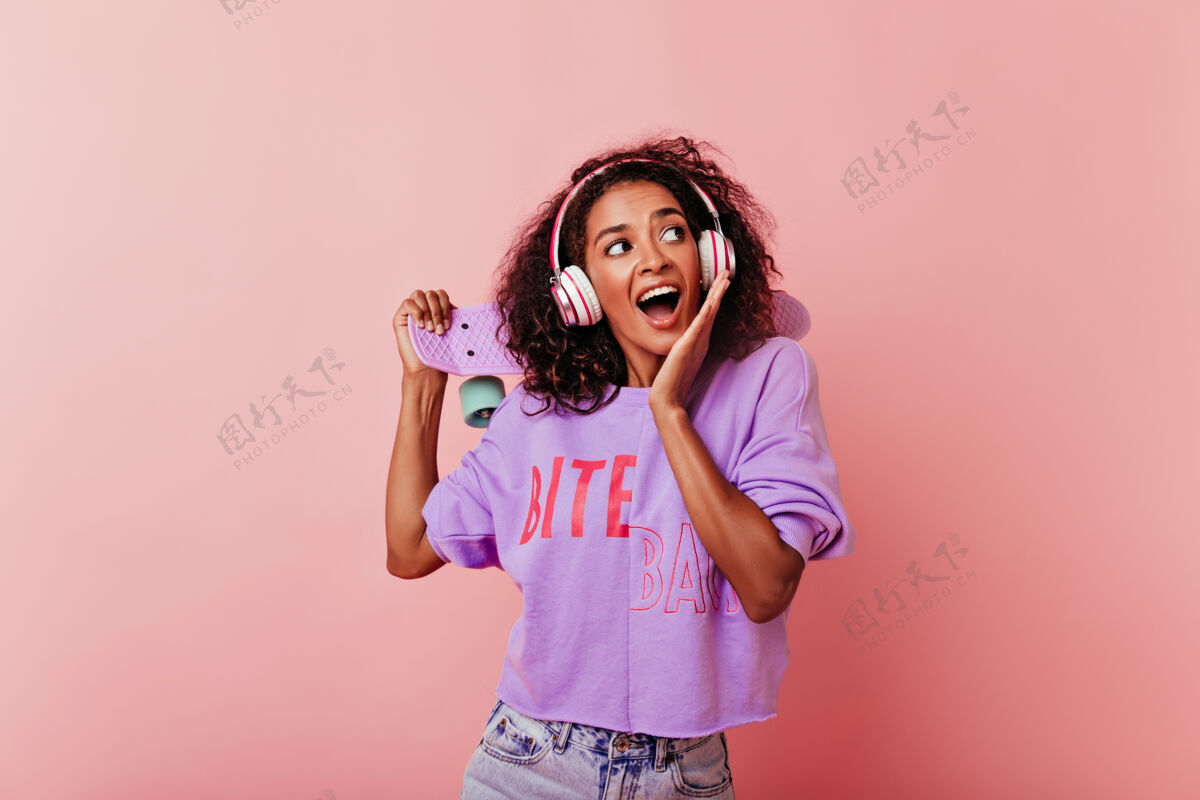 人幸福的黑色卷发女孩与滑板合影幽默的非洲女模特穿着休闲服听音乐模特大笑享受