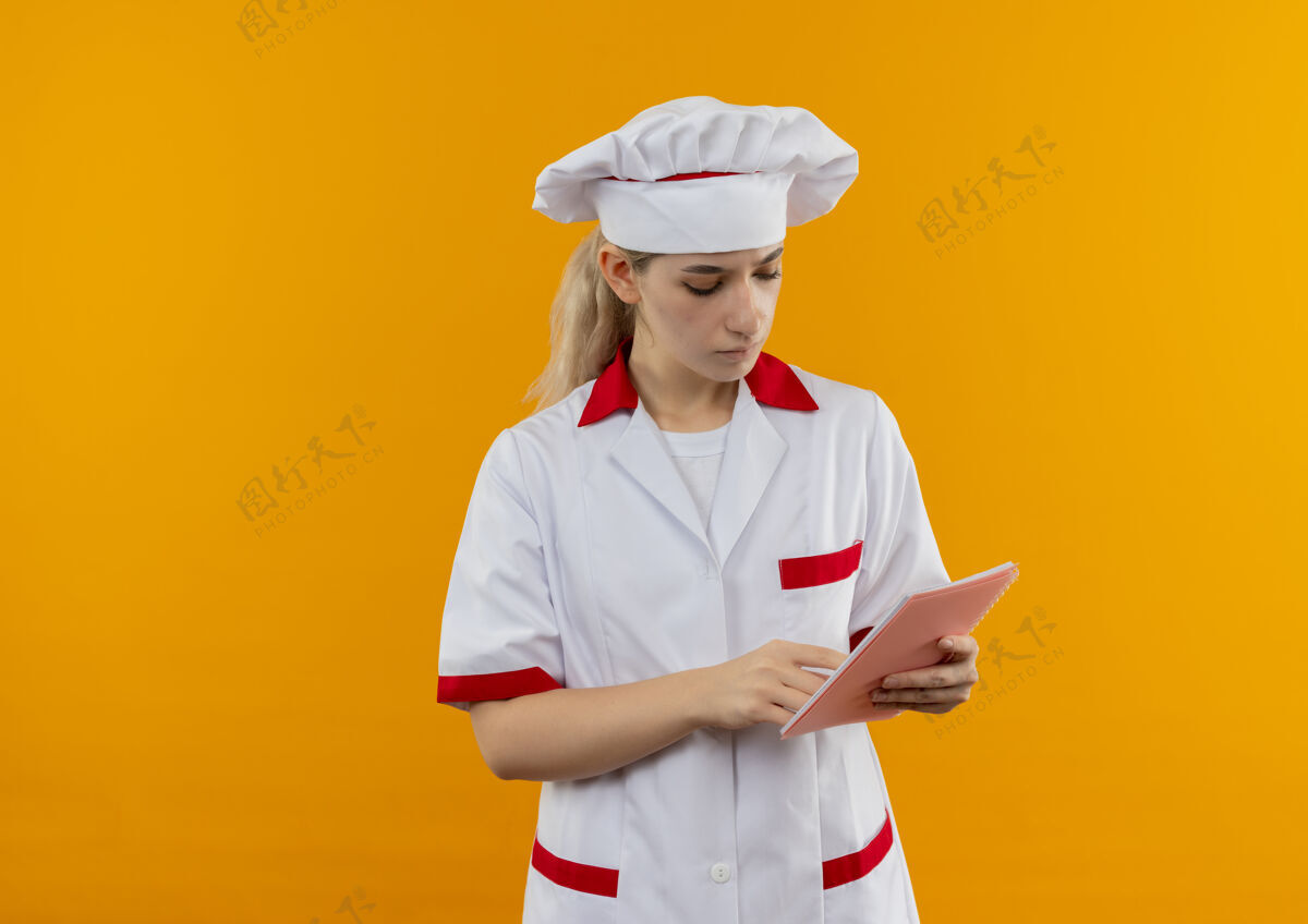 看体贴的年轻漂亮厨师穿着厨师制服 拿着一个手指 把手指放在笔记本上 孤立地放在橙色的空间里空间手指拿着