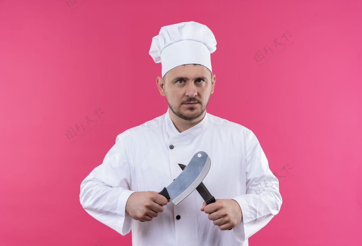 年轻年轻帅气的厨师穿着厨师制服 手里拿着菜刀和刀 隔离在粉红色的空间里严肃粉红刀
