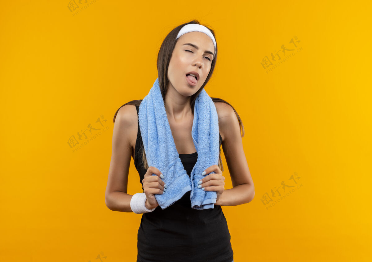 腕带疲惫的年轻漂亮的运动女孩戴着头带和手环拿着毛巾在脖子上展示舌头 一只眼睛闭在橙色的空间头带一个毛巾