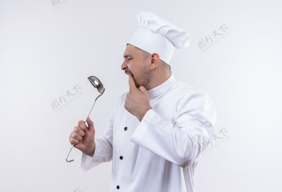 嘴巴年轻帅气的厨师穿着厨师制服 拿着勺子看着它 把手指放在嘴上孤立的空白处年轻拿着看着