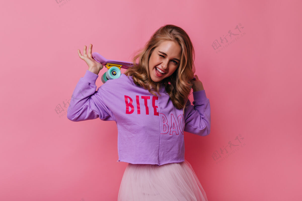 爱好可爱的女孩带着可爱的粉色滑板对着镜头微笑身着紫色衬衫的有趣女模特的肖像运动时尚金发